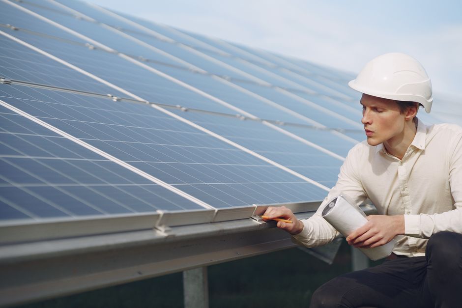 Focused young male employer in formal wear and hardhat working on solar panel station and checking details of modules