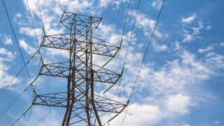 High voltage power line tower framed against a bright blue sky with clouds.