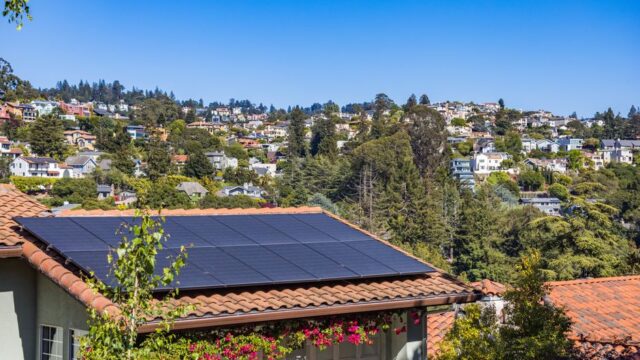 Solar panels on a suburban home, surrounded by lush greenery and a sunny blue sky.
