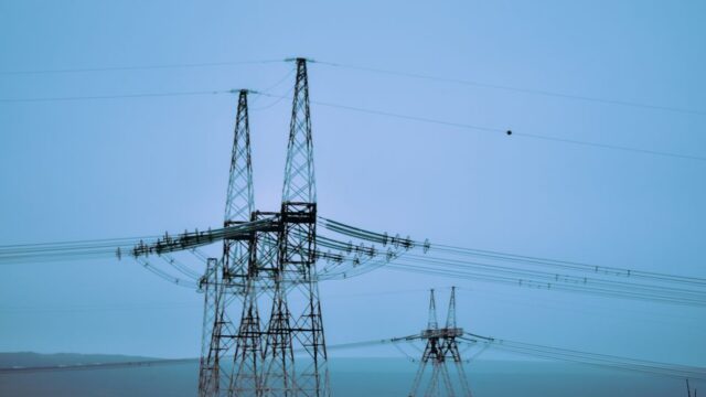 High voltage electricity pylons against ocean with horizon and mountain under blue sky in daytime