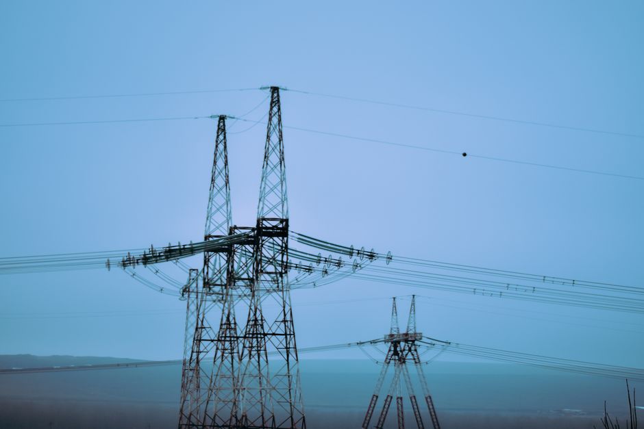 High voltage electricity pylons against ocean with horizon and mountain under blue sky in daytime