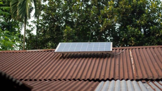 Solar panel on a rusty corrugated roof with green foliage background, symbolizing sustainable energy.
