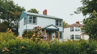 Charming two-story wooden house surrounded by lush greenery and vibrant flowers in a suburban neighborhood.