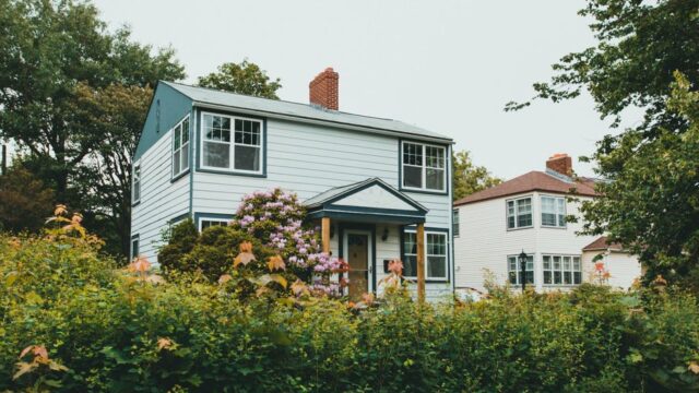 Charming two-story wooden house surrounded by lush greenery and vibrant flowers in a suburban neighborhood.
