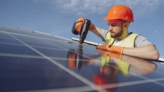 Low angle of serious bearded male electrician in hardhat and protective gloves installing solar photovoltaic panel system using drill