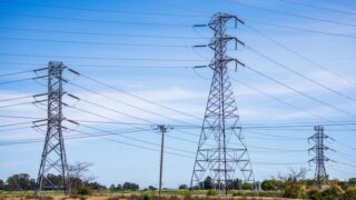 High voltage electric pylons in a field under a clear blue sky demonstrate power transmission infrastructure.