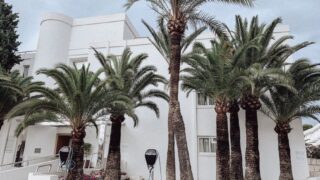 Contemporary white building facade with palm trees and charging stations on a city street.
