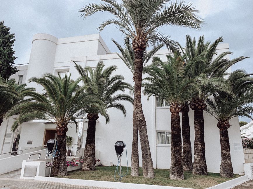 Contemporary white building facade with palm trees and charging stations on a city street.