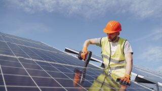 From below of concentrated young bearded male technician in protective clothes mounting solar batteries using drill against blue sky