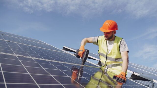 From below of concentrated young bearded male technician in protective clothes mounting solar batteries using drill against blue sky
