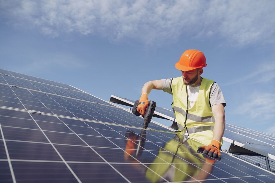 From below of concentrated young bearded male technician in protective clothes mounting solar batteries using drill against blue sky