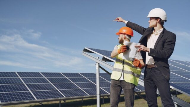 Young businessman in formal outfit and foreman in yellow vest looking away while discussing working process against solar panels