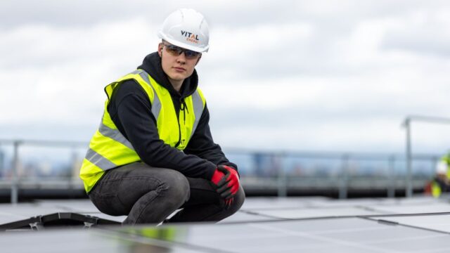 A construction worker inspects rooftop solar panels, wearing safety gear and high visibility vest.