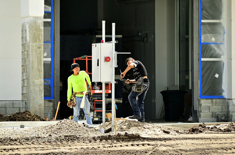 Two construction workers install an electrical panel outside a building site, wearing safety gear.