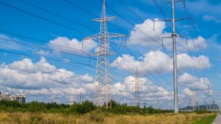 High voltage power lines stretching across a grassy field under bright blue skies with clouds.