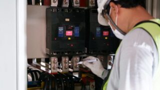 Technician wearing safety gear inspecting electrical panels in an industrial setting.