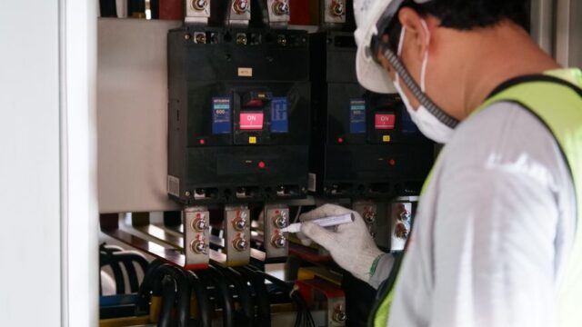 Technician wearing safety gear inspecting electrical panels in an industrial setting.