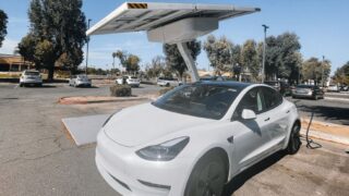 Modern white electric car charging under solar panels in a sunny parking lot.