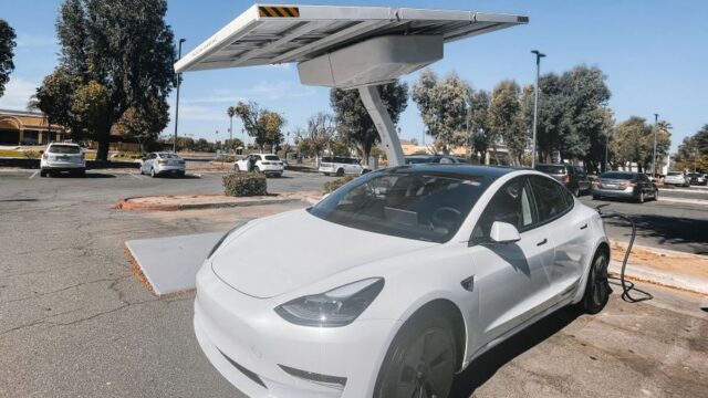 Modern white electric car charging under solar panels in a sunny parking lot.