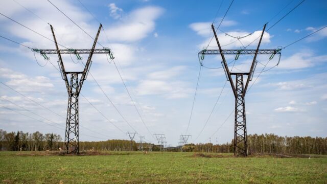 High voltage power lines stretch across a green grass field under a clear blue sky.