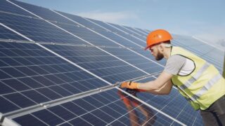 Side view of focused young man in uniform and hardhat checking setup of solar panels