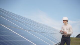 Young serious male engineer in formal outfit and helmet reporting while inspecting solar panel field