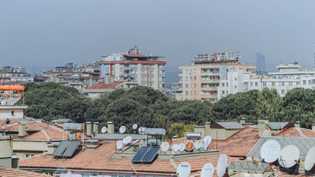 Urban cityscape featuring rooftops with solar panels and satellite dishes surrounded by buildings and trees.