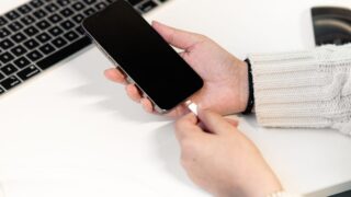 A woman plugs her smartphone into a charger at a desk, next to a laptop, illustrating modern technology usage.