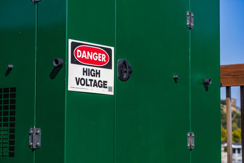 Green electrical box with warning 'Danger High Voltage' sign under clear sky.