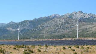 A scenic view of windmills and solar panels against mountainous terrain under a clear blue sky.
