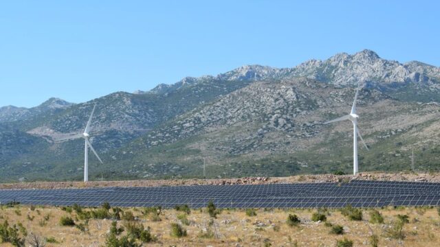 A scenic view of windmills and solar panels against mountainous terrain under a clear blue sky.