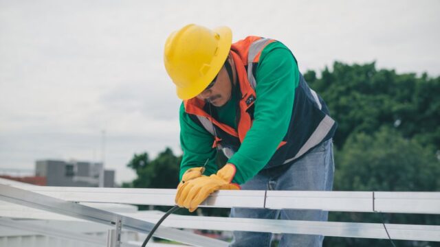 A construction worker with safety gear installing solar panels on a sunny day.