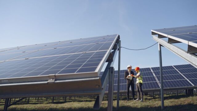 Supervisor in hardhat and formal wear discussing project with workman in uniform while standing with papers near modern solar panels under blue sky