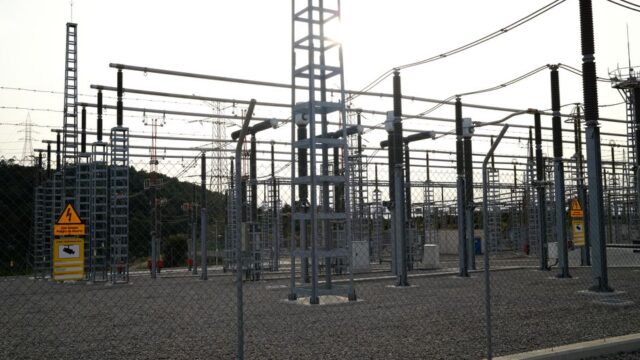 Outdoor electrical power substation with high voltage equipment and safety signs, surrounded by fencing.