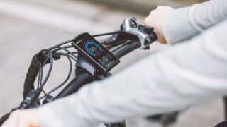 Hands gripping the handlebar of an electric bicycle, showcasing the digital display screen for speed and battery status.
