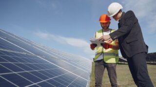 Low angle of young inspector and foreman in hardhats checking documentation against modern solar panels in field
