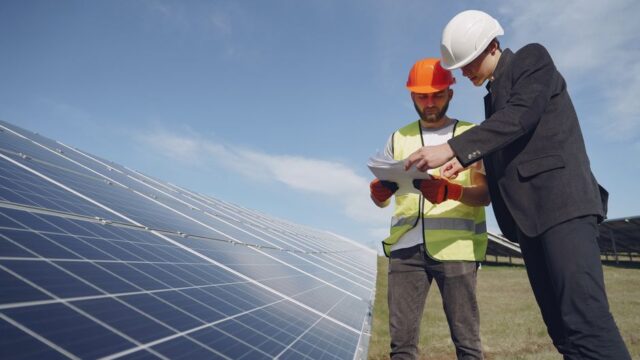Low angle of young inspector and foreman in hardhats checking documentation against modern solar panels in field