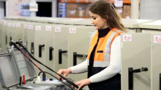 A female electrician in safety gear checks electrical panels indoors.