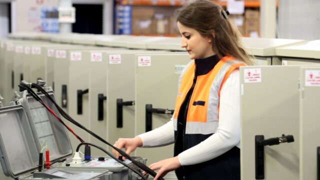A female electrician in safety gear checks electrical panels indoors.