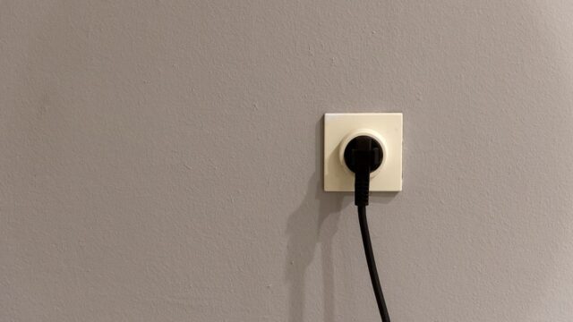 A black plug inserted into a white wall socket on a plain grey wall.