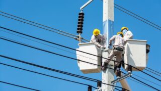 Electricians wearing safety gear work on power lines using lifts against a clear blue sky.