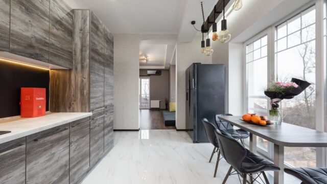 Table with fruits and flowers placed near window near counter and gray cupboards in stylish kitchen with fridge in apartment