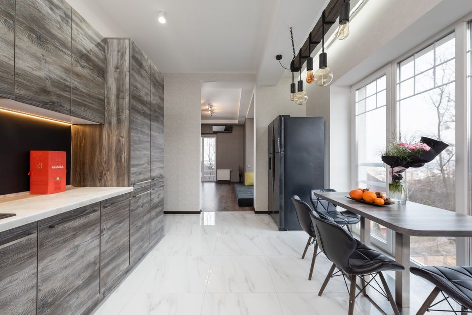 Table with fruits and flowers placed near window near counter and gray cupboards in stylish kitchen with fridge in apartment
