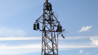 A high voltage power line tower set against a clear blue sky with visible clouds.