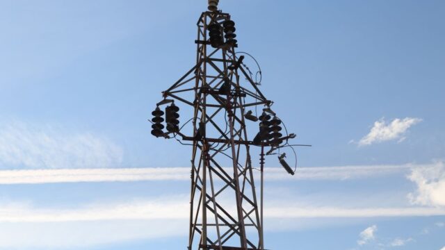 A high voltage power line tower set against a clear blue sky with visible clouds.