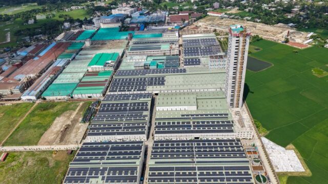 Sprawling industrial complex with solar panels and green fields, seen from above.