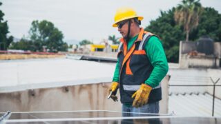 A solar technician in protective gear inspecting rooftop solar panels outdoors.