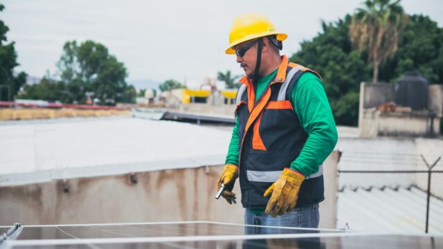 A solar technician in protective gear inspecting rooftop solar panels outdoors.