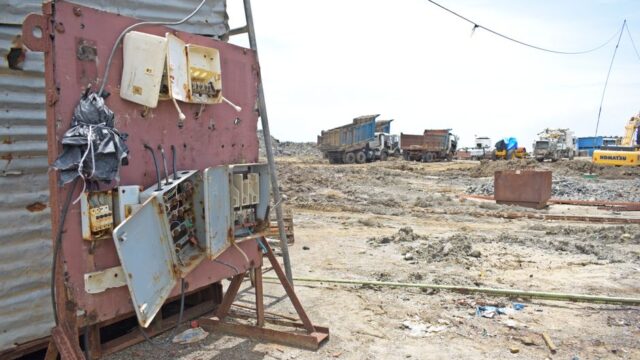 A barren construction site with rusted electrical panel and heavy equipment in the background.