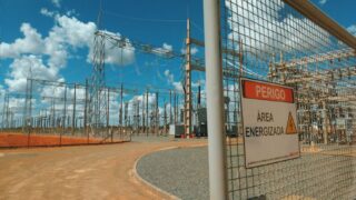 Empty high voltage station with wires and pillars located in sunny sandy area behind metal fence with warning sign